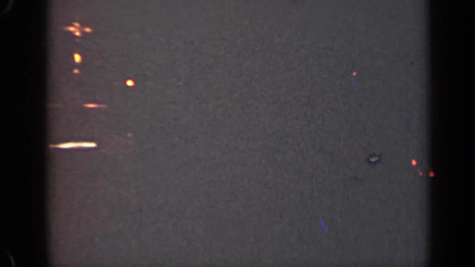 1947 Illinois 8mm frame showing dim, out-of-focus orange lights, dark film edges, grain, and a small bright horizontal streak.