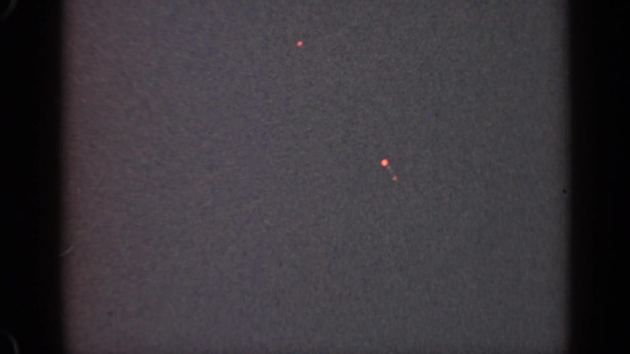Dark grainy frame with black film borders and a few small red-orange light points.