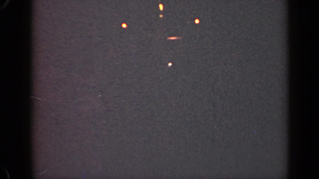 Dark grainy frame with several orange lights, a pale streak, and black film borders.