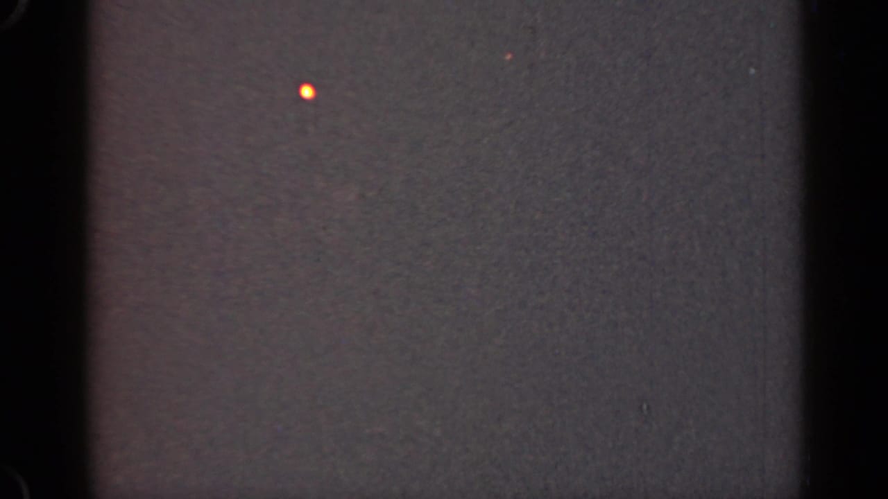 Dark grainy frame with black film edge, faint orange dot, dust, and muted gray texture.