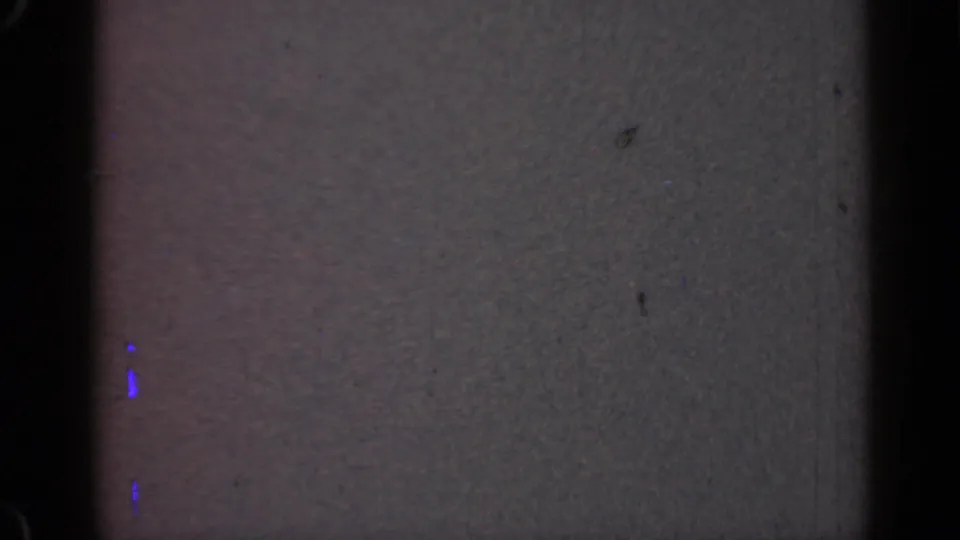 1947 Illinois 8mm frame showing a muted gray field with heavy grain, dark vertical bands, small dust spots, and faint blue marks along the lower left edge.