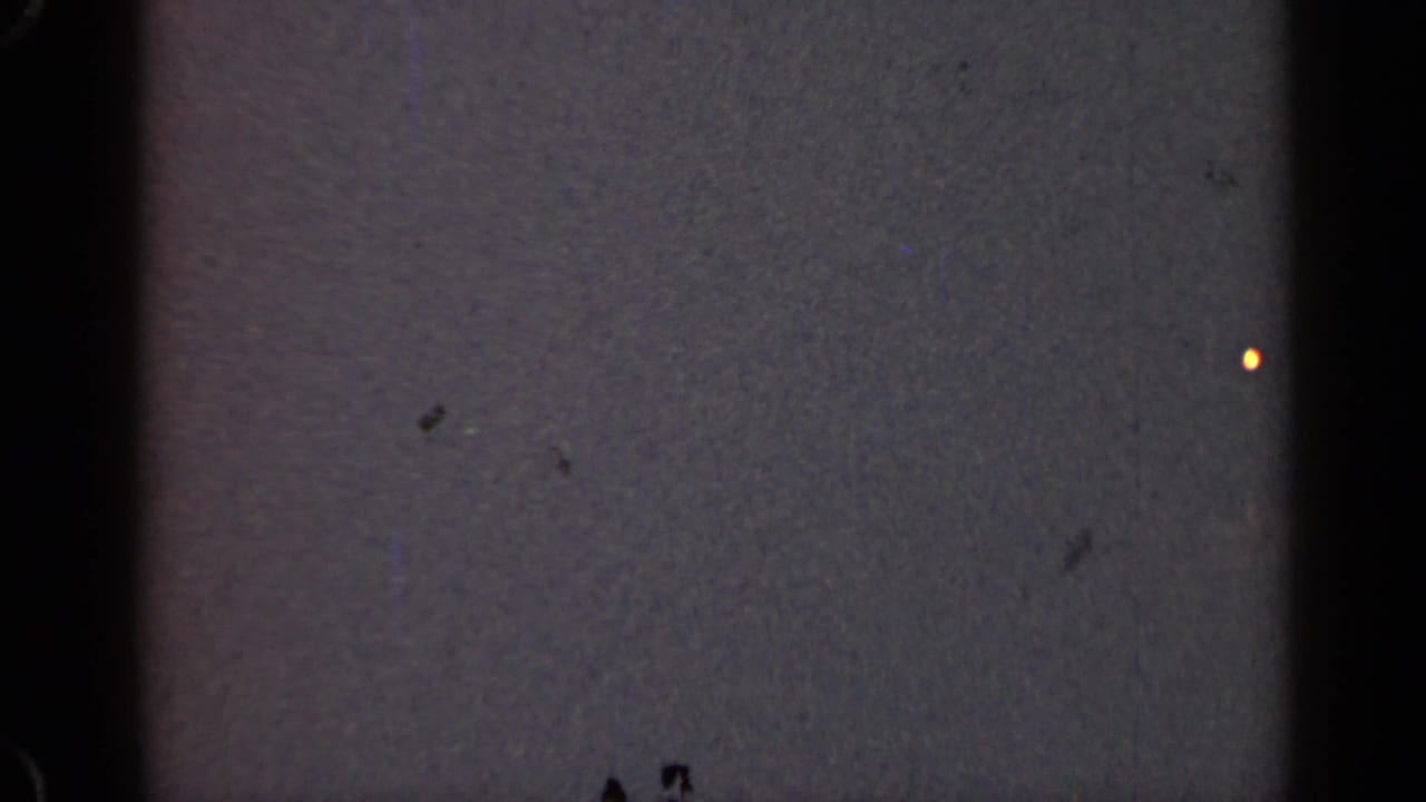 Dark, grainy film frame with gate edges and only a tiny warm light visible.