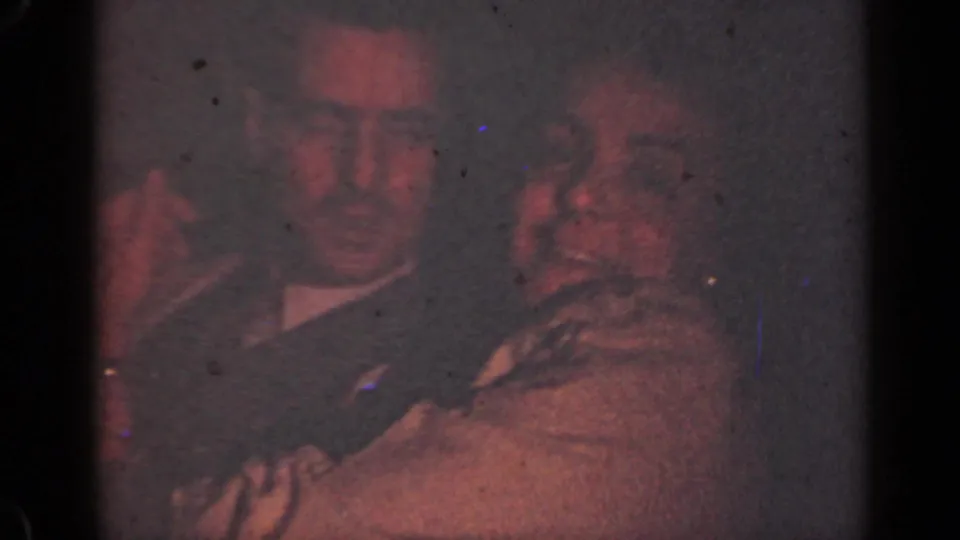 1947 Illinois 8mm frame of an adult couple leaning close in a dim room, with faces softly lit by reddish light.