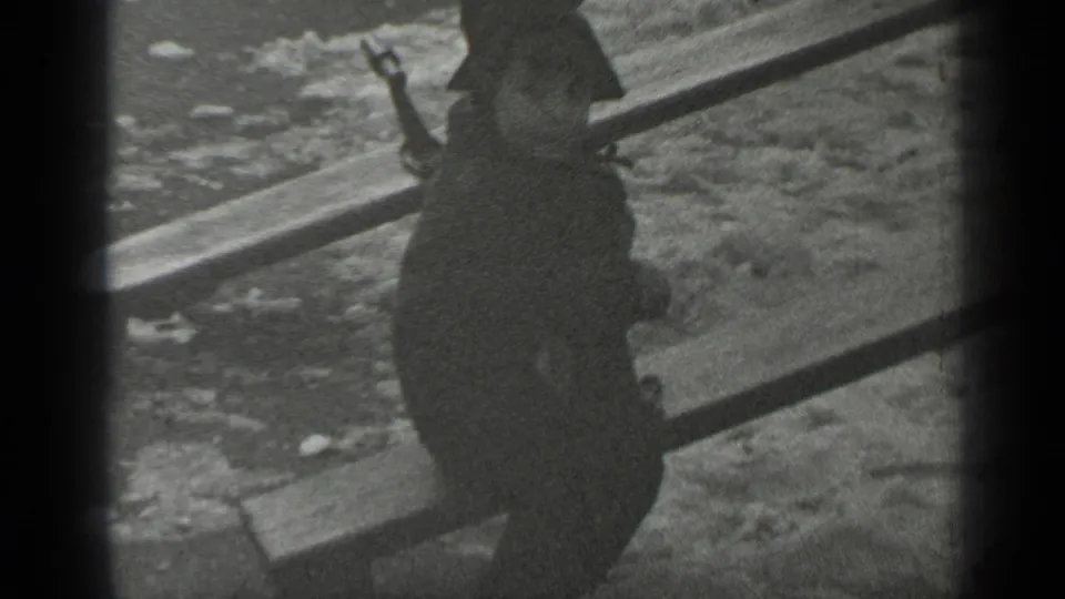 The child, seen mostly from behind in a dark coat and hat, sits on the seesaw above snowy park ground.