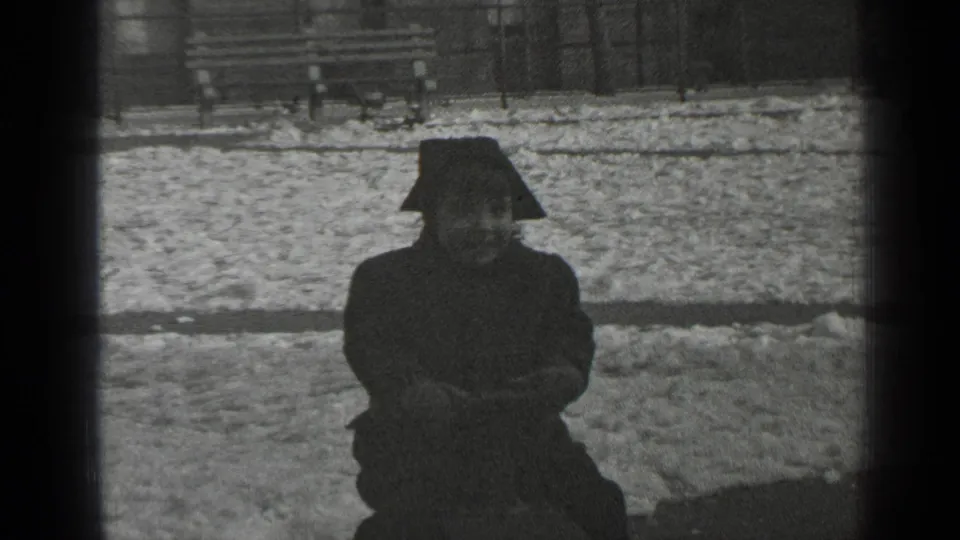 The child sits on the seesaw in a dark winter coat, with snow, a bench, and fencing visible behind them.