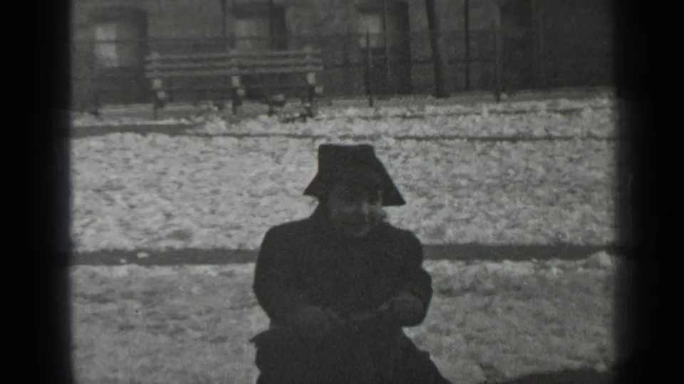 In 1947 New York footage, a bundled child sits low on a seesaw with snow, a bench, and railings behind them.