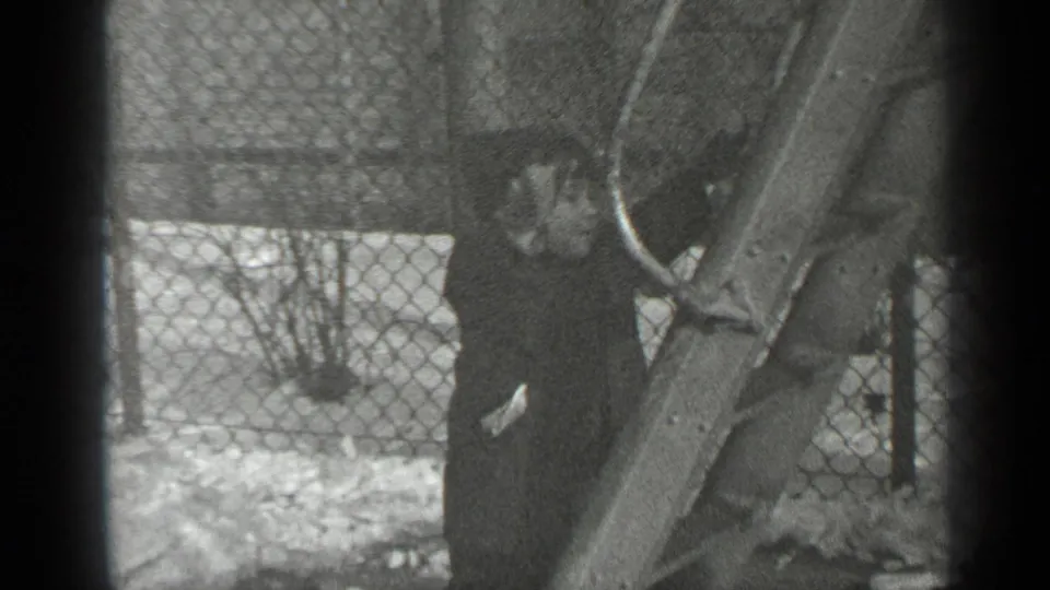 In 1947 New York home-movie footage, a small child in a dark coat and hat climbs a wooden playground slide beside a snowy fence.