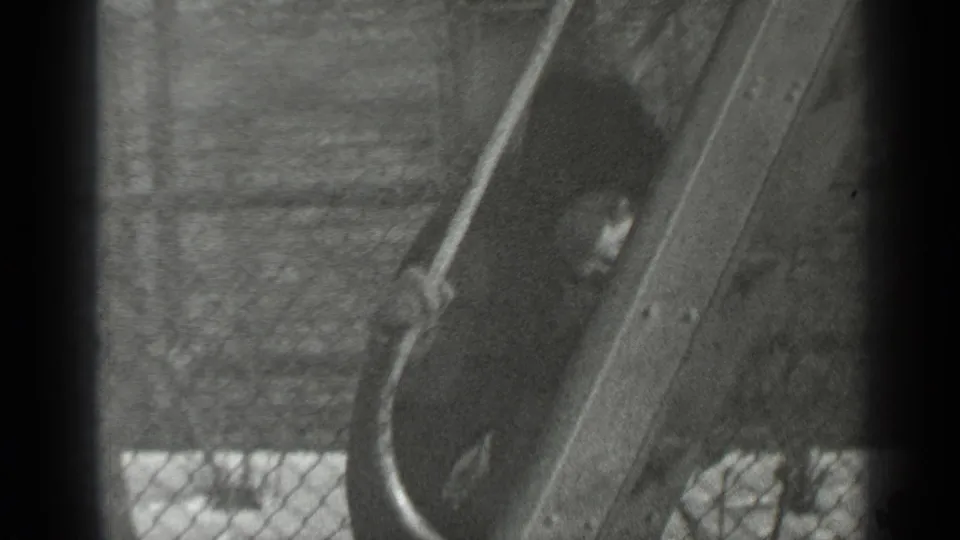The child is mostly obscured by the slide as the black-and-white 8mm frame shows snow, fence, and playground structure.