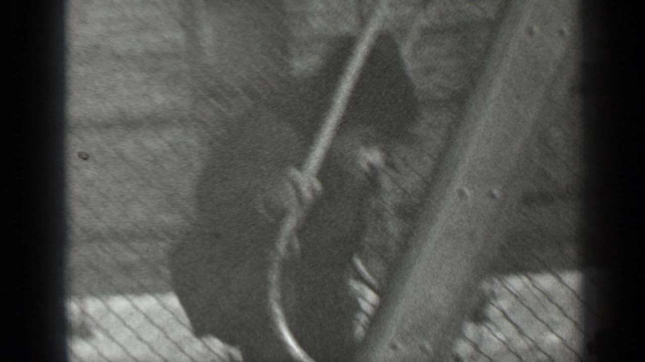In black-and-white 1947 New York home-movie footage, a young boy climbs near a wooden play structure seen through chain-link fencing.