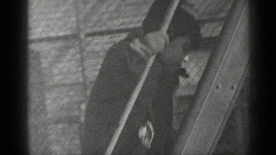 The boy is partly obscured by fencing and frame edges as he climbs beside a wooden structure against a brick wall.