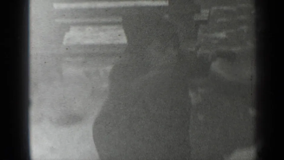 Black-and-white 8mm frame showing a dark-clad person passing near wooden rails or stall-like structures in a public setting.
