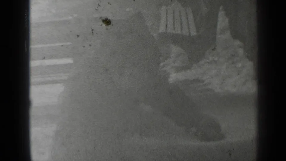 Black-and-white 8mm frame showing a pedestrian's lower body and shoes beside wooden rails or stall edges.