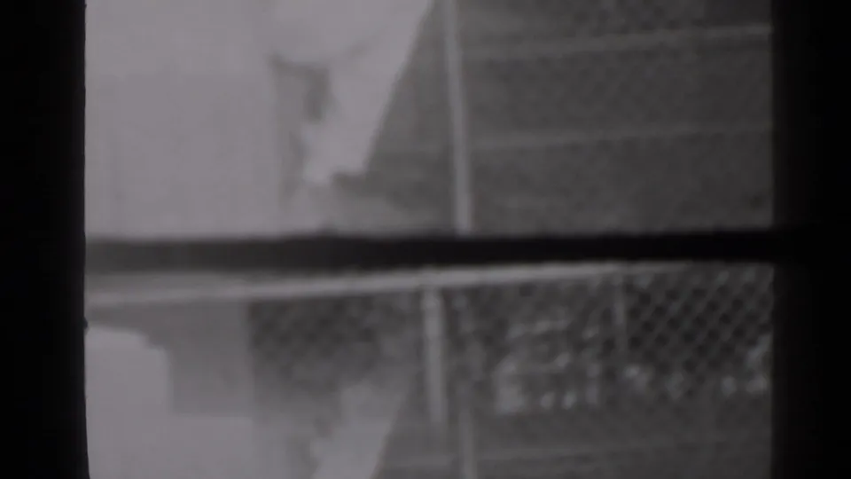 Soft 1947 New York 8mm frame showing a chain-link fence behind dark vertical window or wall divisions.