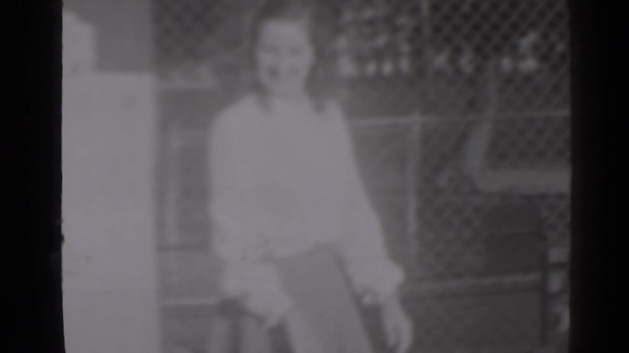 Woman in a light blouse stands near a wall with a chain-link fence behind her.
