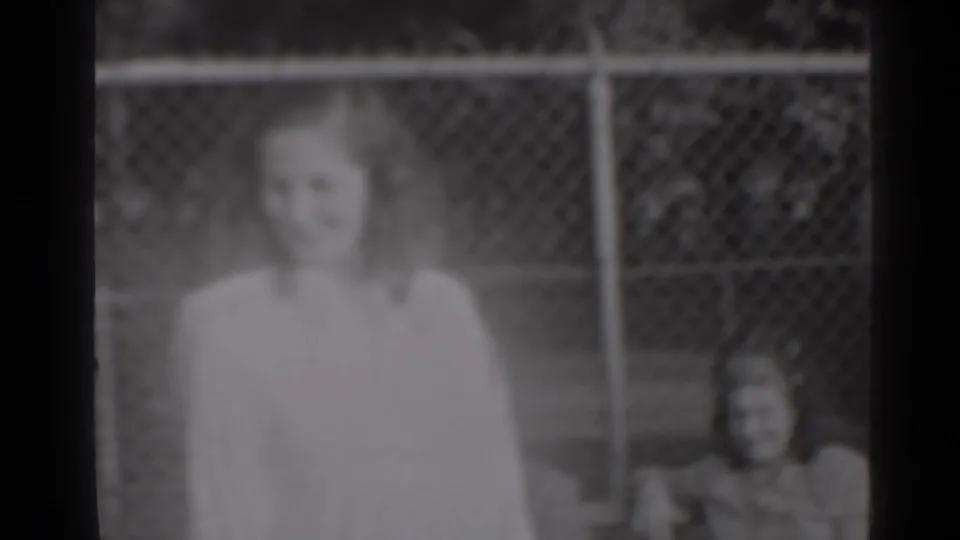 The woman faces the camera in soft black-and-white 8mm footage, with a chain-link fence and background figure visible.