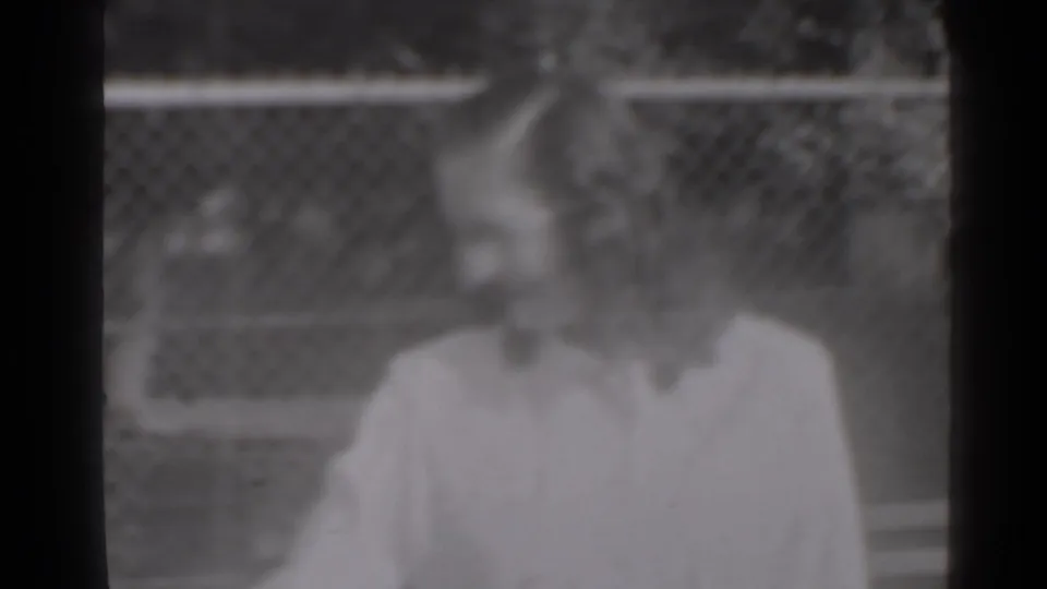 The woman remains near the fence in a soft 1947 New York home-movie frame, with other people faintly visible behind her.