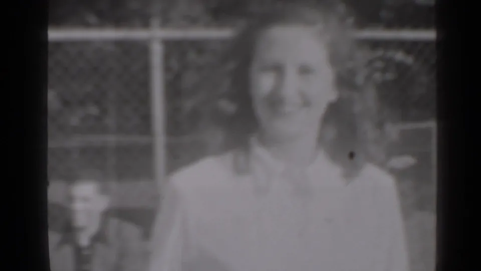 In 1947 New York home-movie footage, a smiling woman faces the camera outdoors near a chain-link fence.