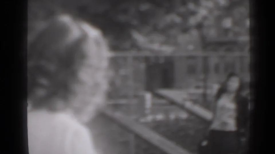 The camera looks past a woman in the foreground toward a man standing near outdoor railings in New York home-movie footage.