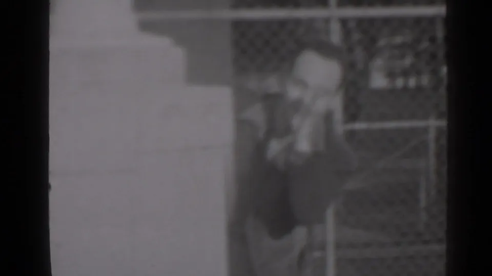 A man stands beside a chain-link fence in a soft 1947 New York home-movie frame, partly obscured by dark framing.