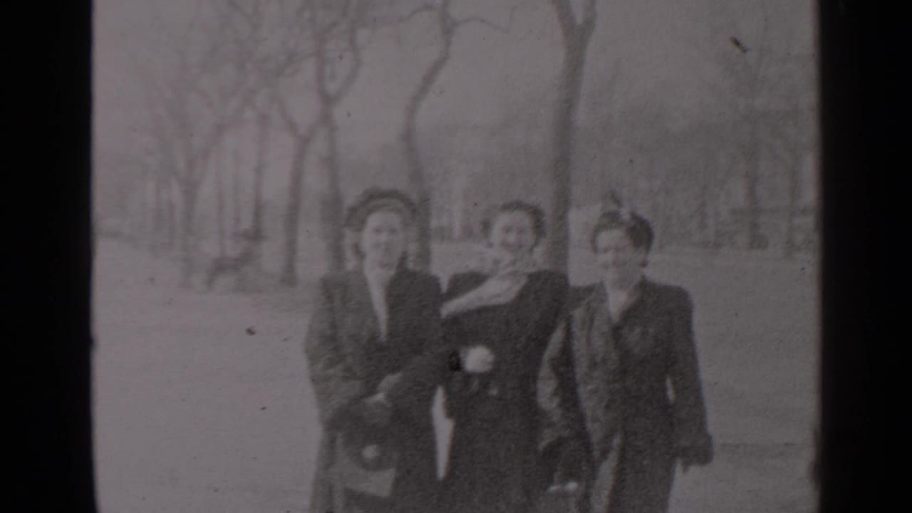 Three women in dark coats walk toward the camera on a bare-tree park path.