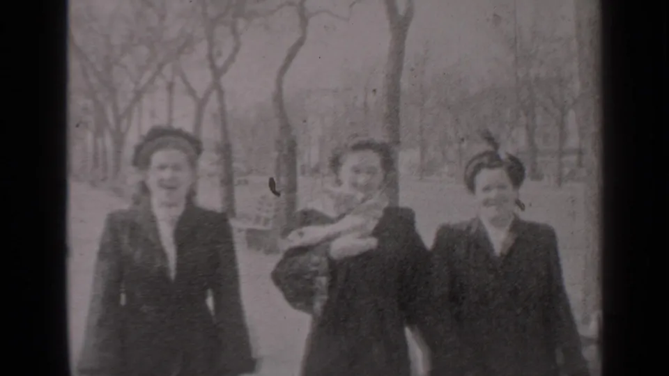 Three adult women in coats and hats approach the camera in a New York park-like area, with one managing a scarf in the wind.