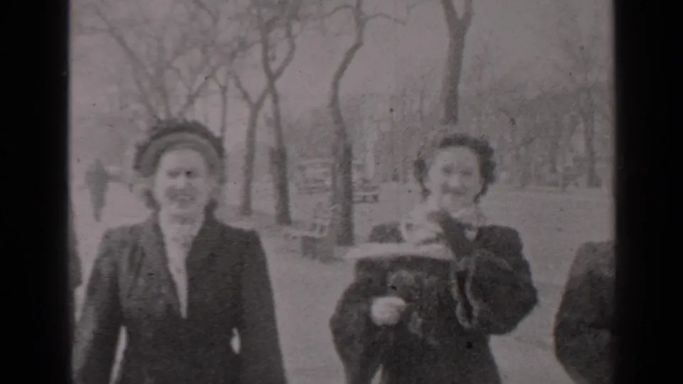 Two of the women fill the frame as the group passes close to the camera, with the scarf still caught by the wind in the park setting.