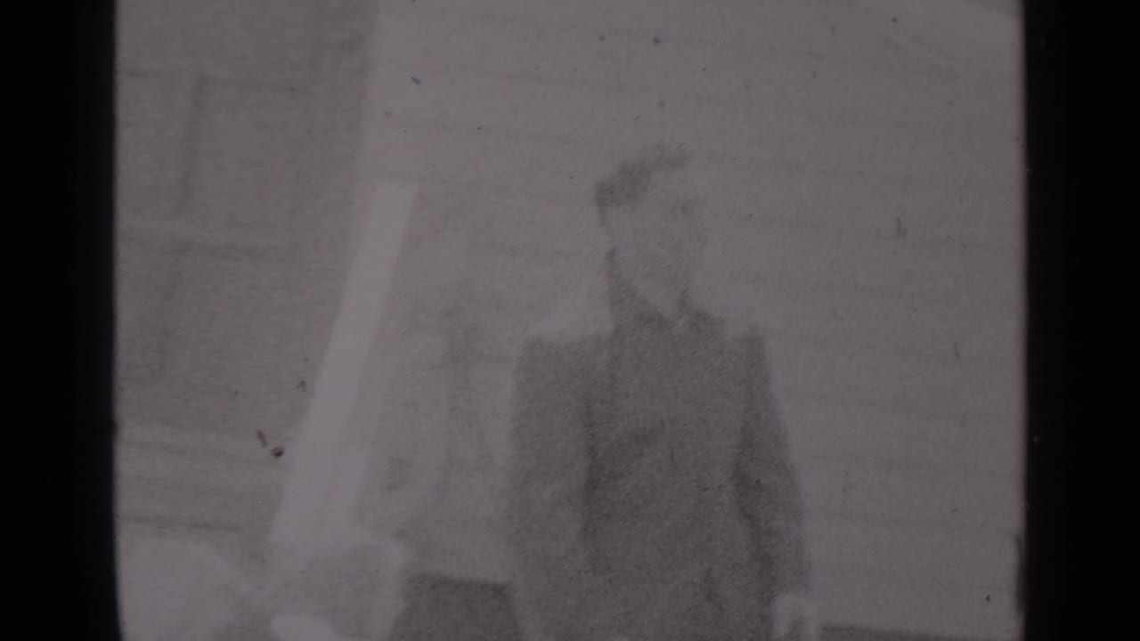 Two men standing outdoors near clapboard siding, with one figure partly blurred.