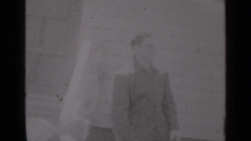 Two men in 1947 New York home-movie footage stand outside a clapboard house, one partly obscured by soft focus and film grain.