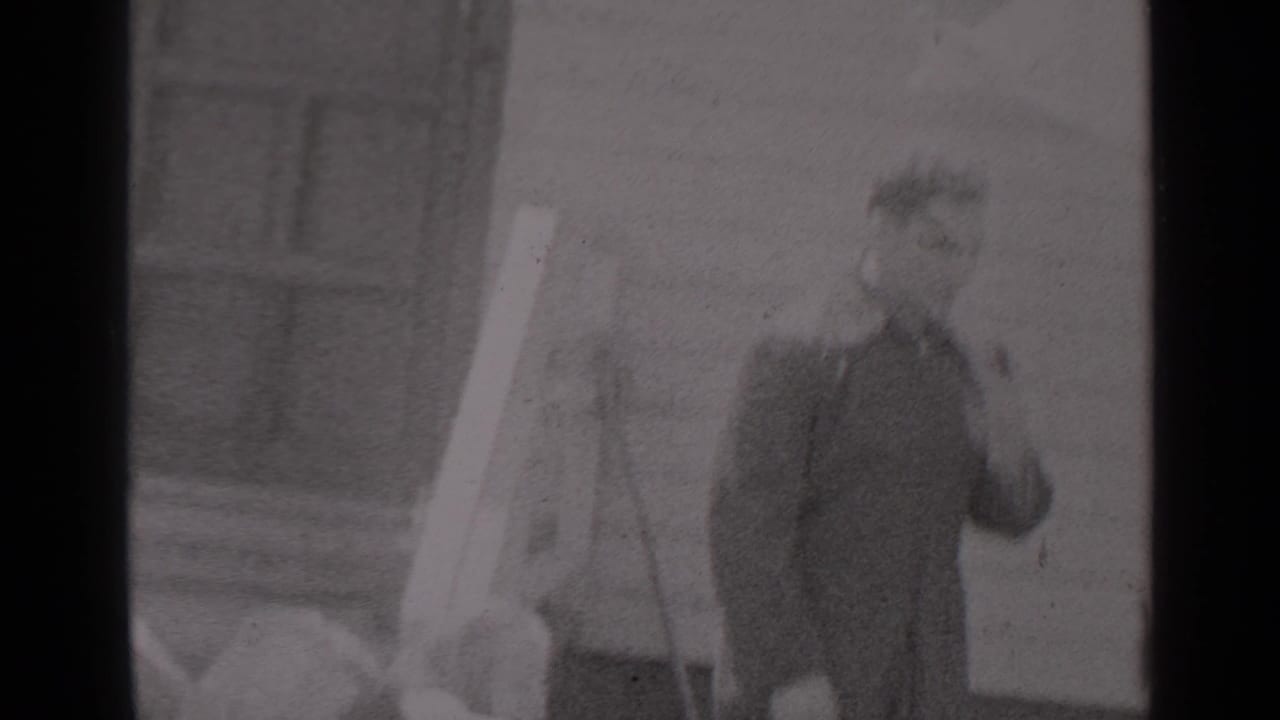 Two men in uniform or dark outerwear stand outside a New York house in 1947, with one gesturing beside a pale hanging fabric or pole.