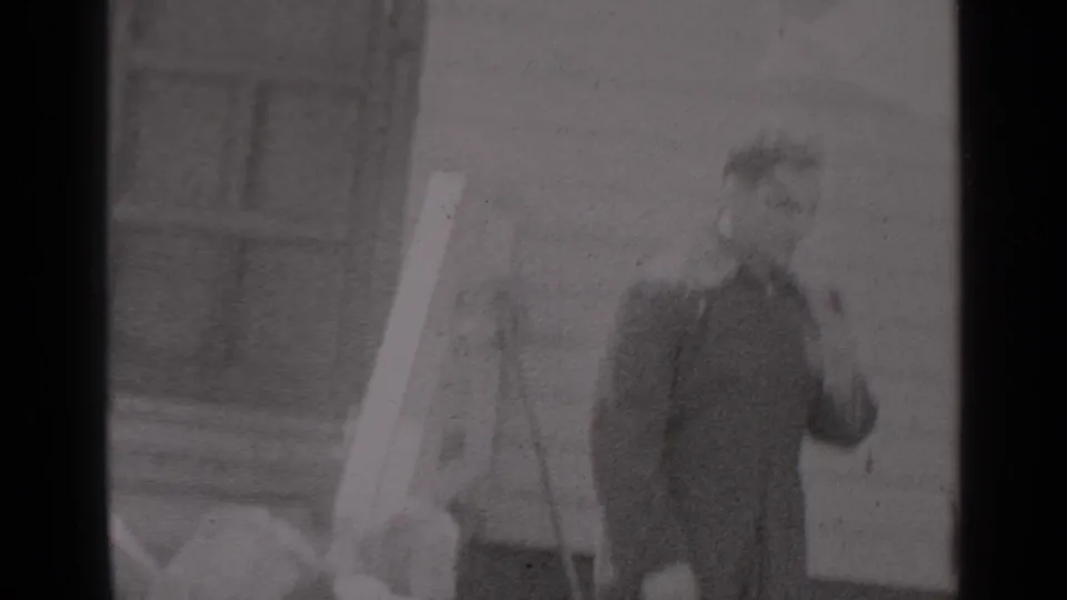 Two men in uniform or dark outerwear stand outside a New York house in 1947, with one gesturing beside a pale hanging fabric or pole.