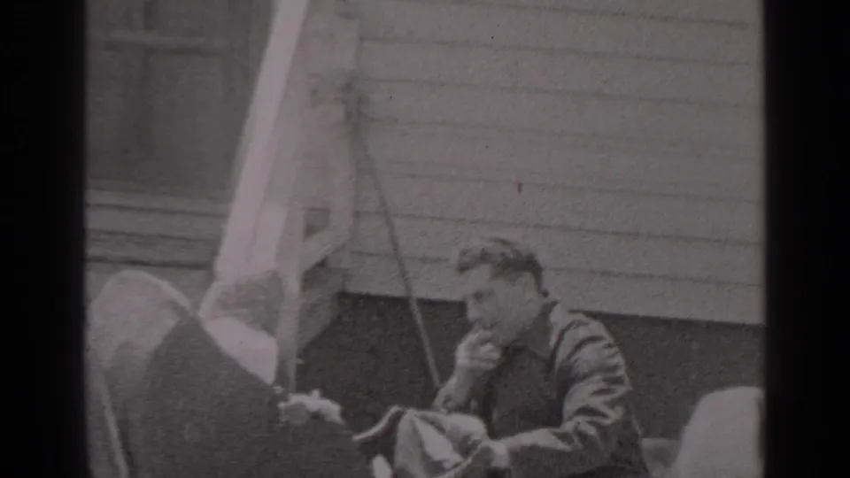 A man in dark clothing smokes a cigarette outside a clapboard house in 1947 New York home-movie footage, with foreground figures partly visible.
