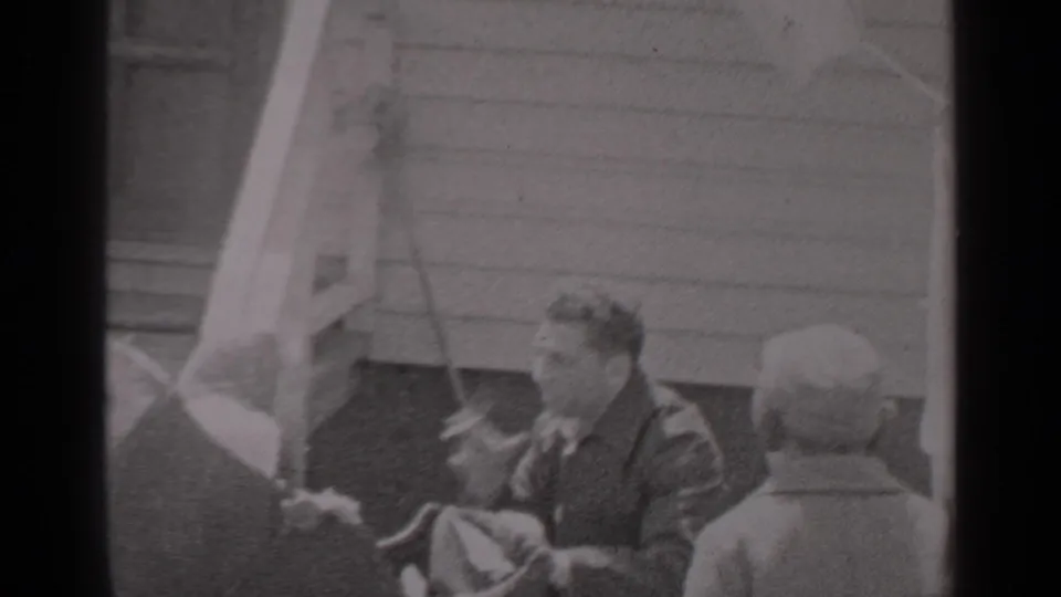 A man smokes a cigarette outside a New York clapboard house in 1947, framed by foreground figures and visible 8mm grain.
