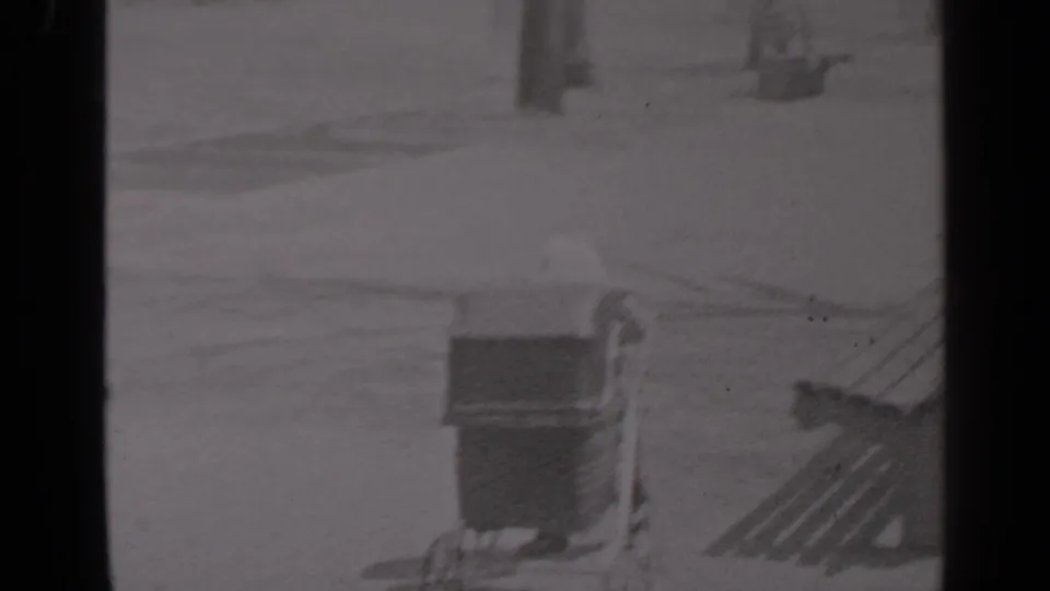 Black-and-white 8mm frame of a vintage baby carriage moving past park benches on a bright snow-covered path.