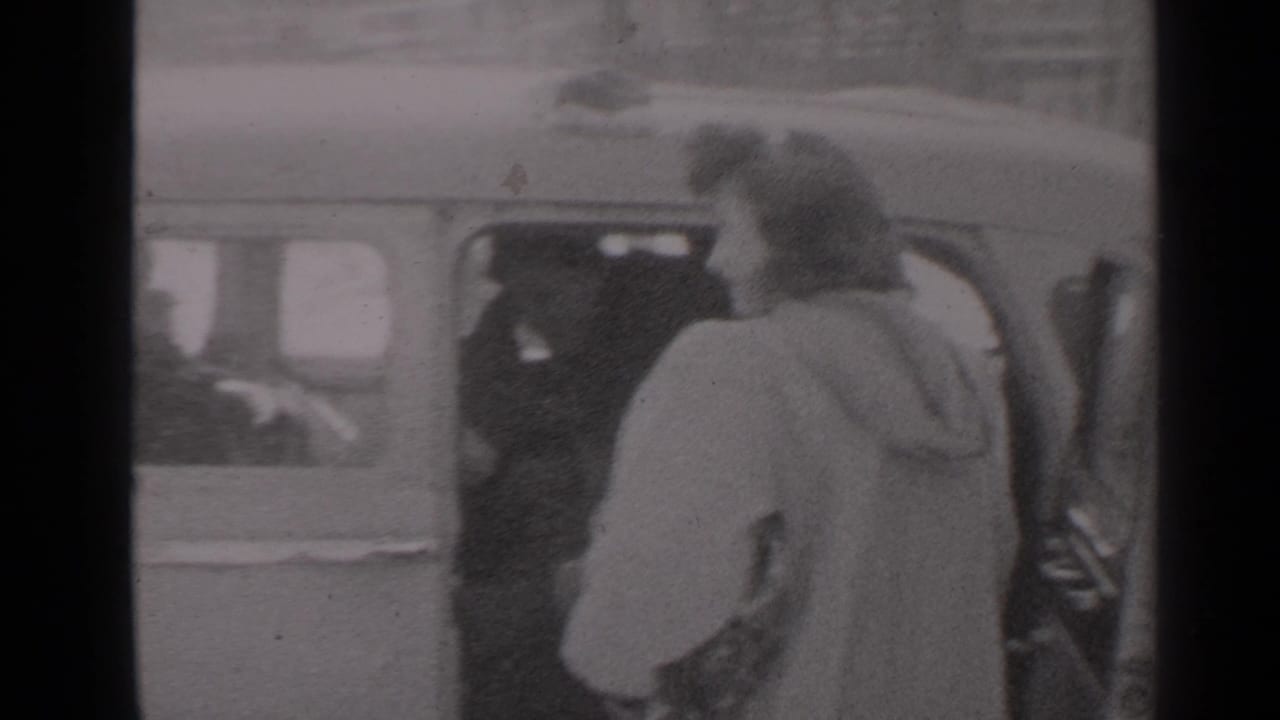 Woman in a light coat stands beside an open car door on a city street.