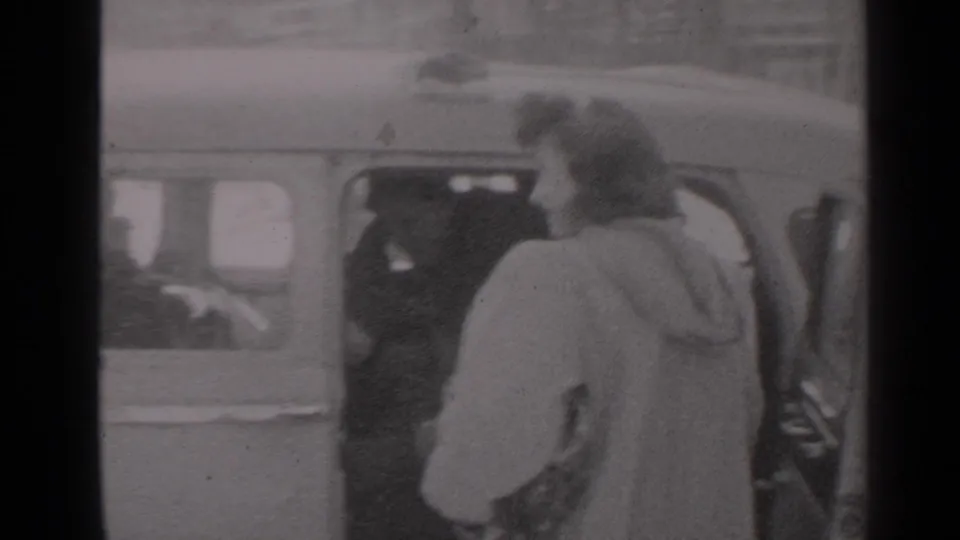 1947 New York frame with a woman in a light coat at an open car door, seen in soft black-and-white 8mm footage.