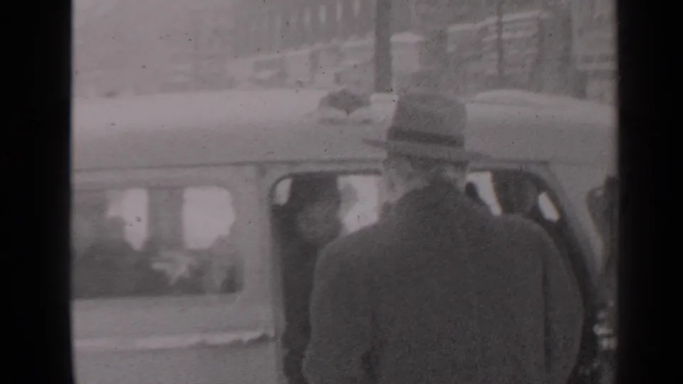In a 1947 New York home-movie frame, bundled adults appear beside a car near a snowy residential entrance.