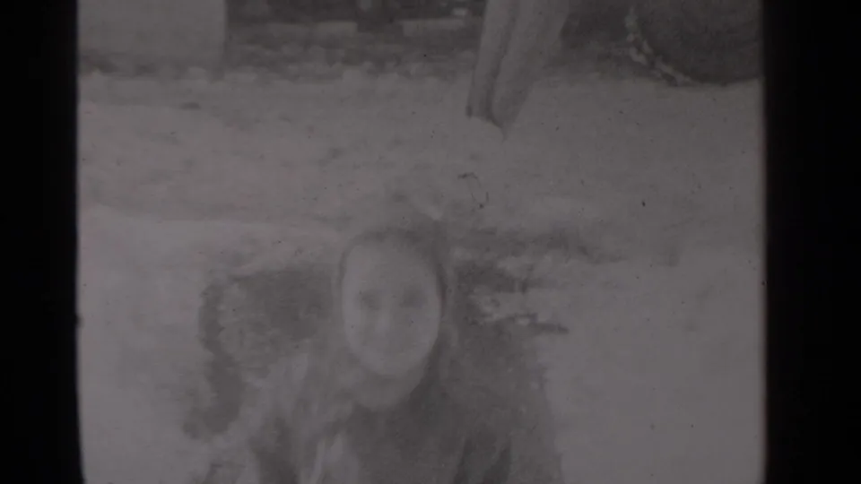 A young child appears in winter clothing near a snowy residential porch in the 1947 New York home-movie setting.