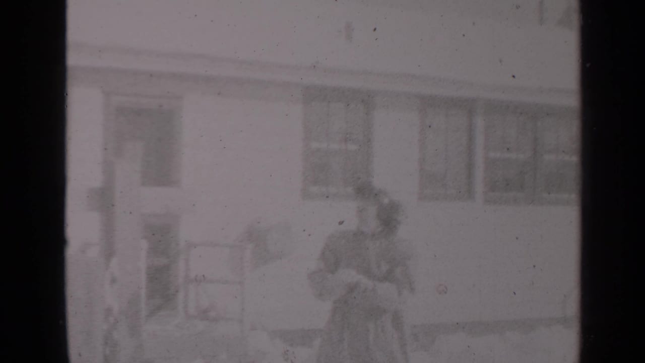 Woman in winter clothing stands in snowy yard near a house while holding a small child.
