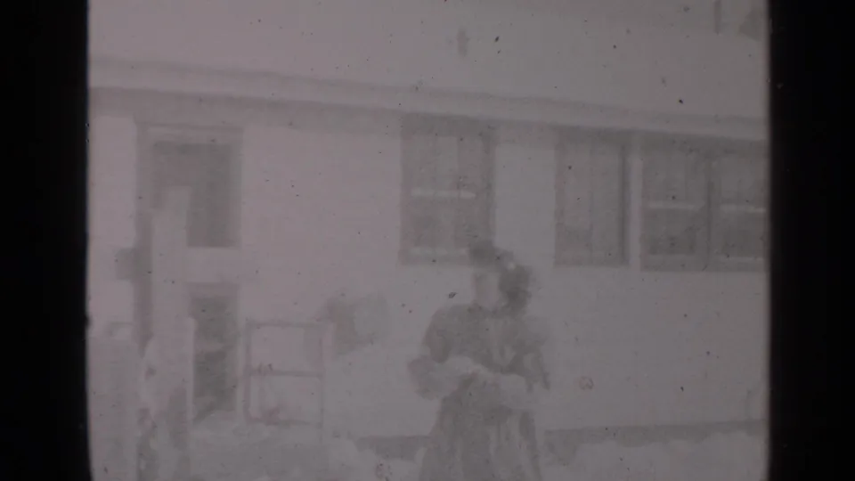 1947 New York home-movie frame showing a woman in winter clothing with a small child outside a snowy residential house.