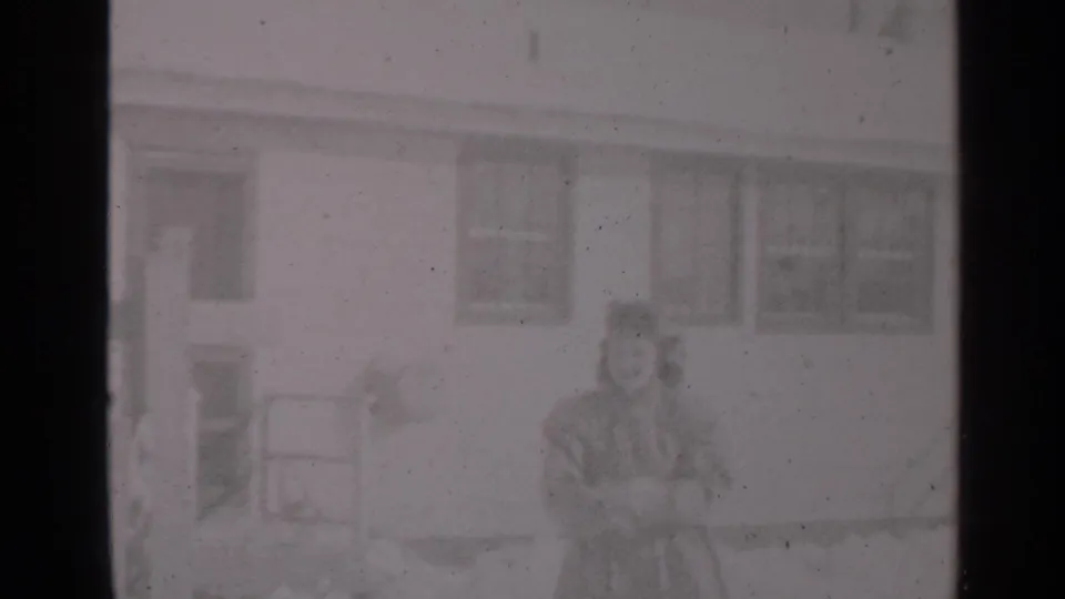 Soft 8mm frame from a 1947 New York winter home movie, with a woman and small child outside a residential building.