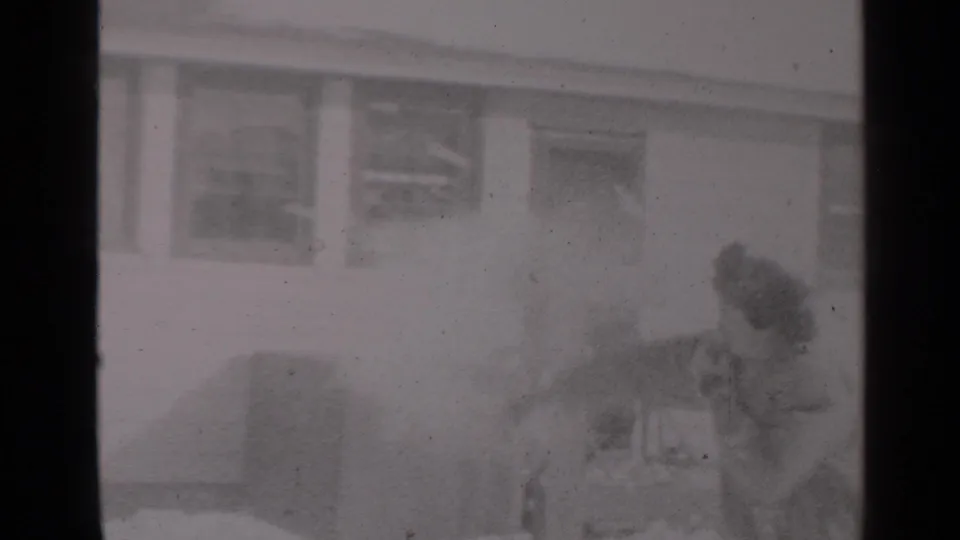 1947 New York home-movie frame showing a man with a shovel near a woman on snowy ground by a residential building.