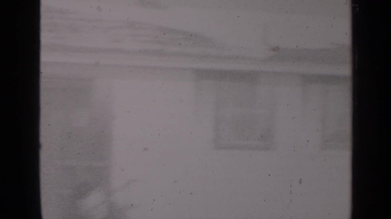 Snowy exterior seen through dark window framing, with a house facade faintly visible.