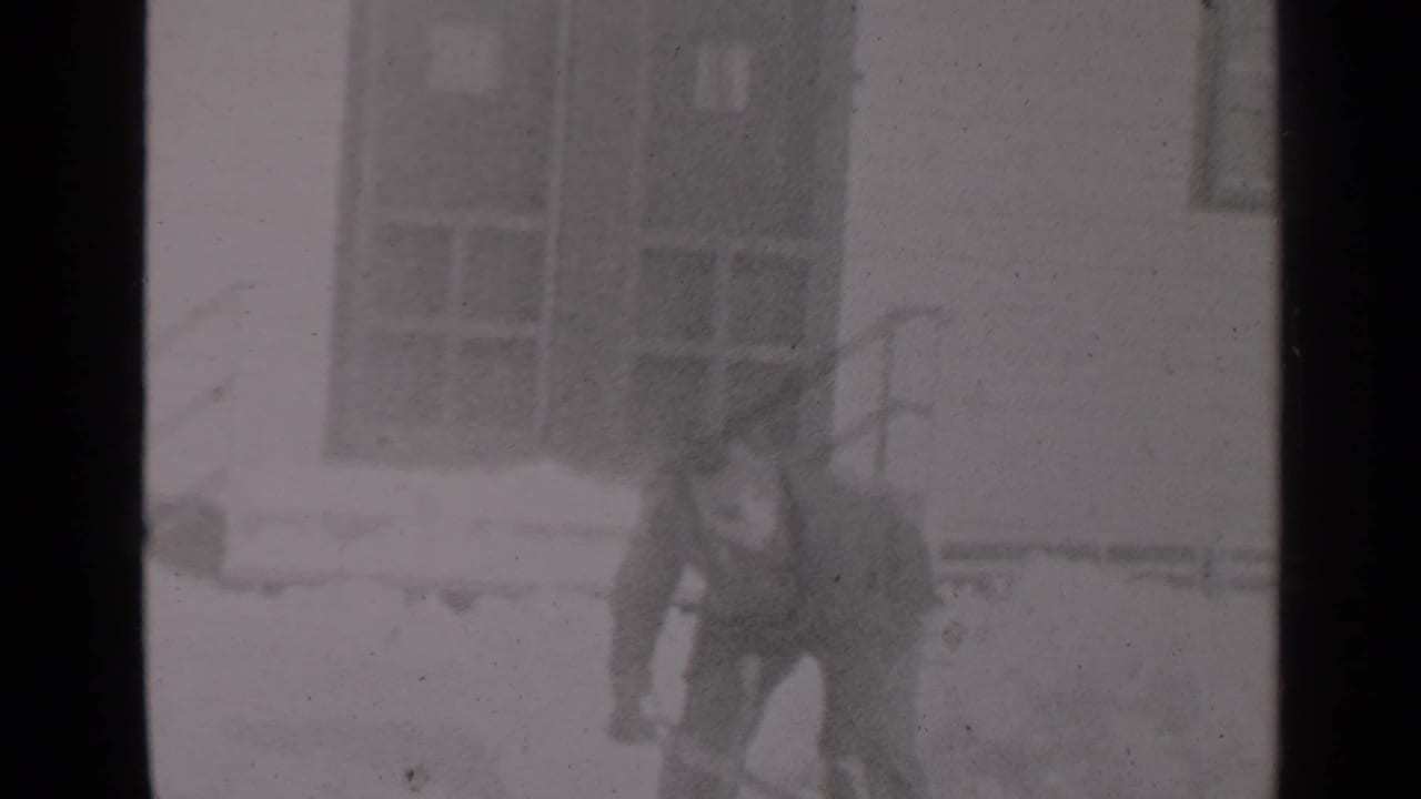 Bundled man bends with a shovel near a snowy house exterior.