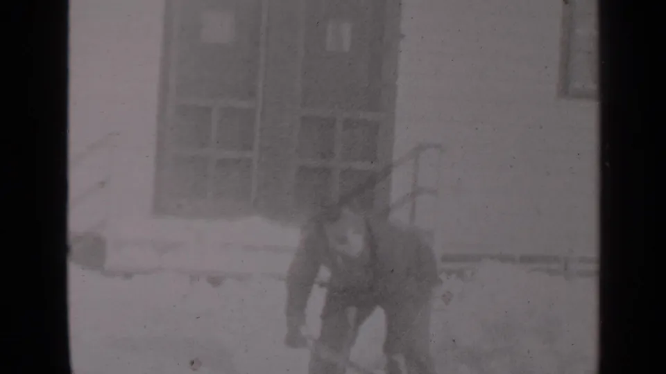 A bundled adult man shovels snow outside a New York house in this soft black-and-white 1947 home-movie frame.