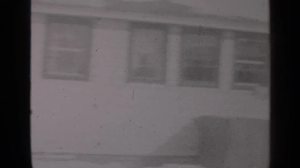 The frame shows a snowy residential facade in New York, partly blocked by a dark vertical window frame and softened by 8mm grain.