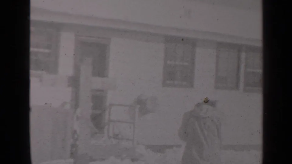 A bundled man stands with a shovel near porch steps outside a snow-covered New York residence in a 1947 home-movie frame.