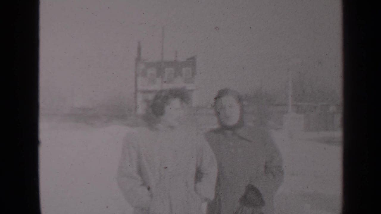 Two women in winter coats walk through a snowy street with buildings behind them.