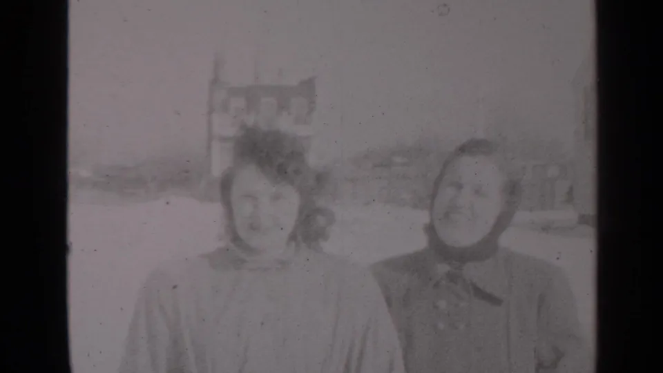 A closer 1947 New York home-movie frame shows two women smiling in winter coats with snow and buildings behind them.