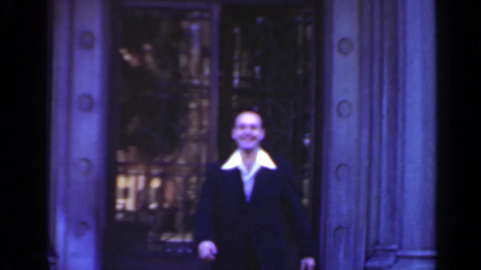 The man in a dark suit and white collar is framed closer at the stone doorway, with soft focus and blue-purple color shift.