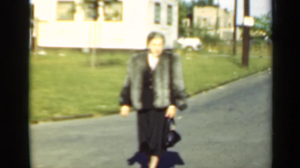 A woman in a fur coat and dark dress walks toward the camera on a residential street, carrying a handbag in 1947 New York footage.